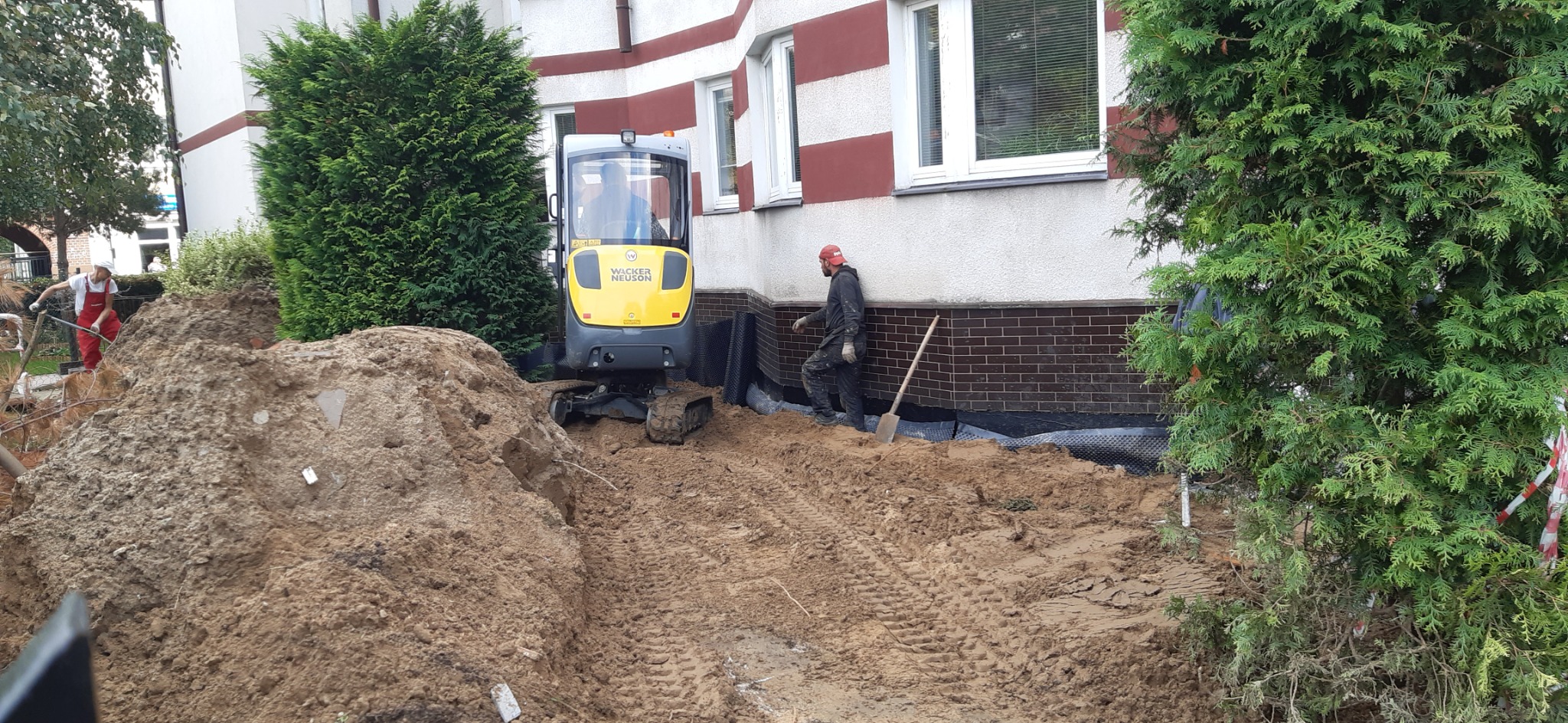 Żółta mini koparka Wacker Neuson podczas prac ziemnych przy izolacji fundamentów budynku z ceglaną podmurówką, widoczni dwaj pracownicy.