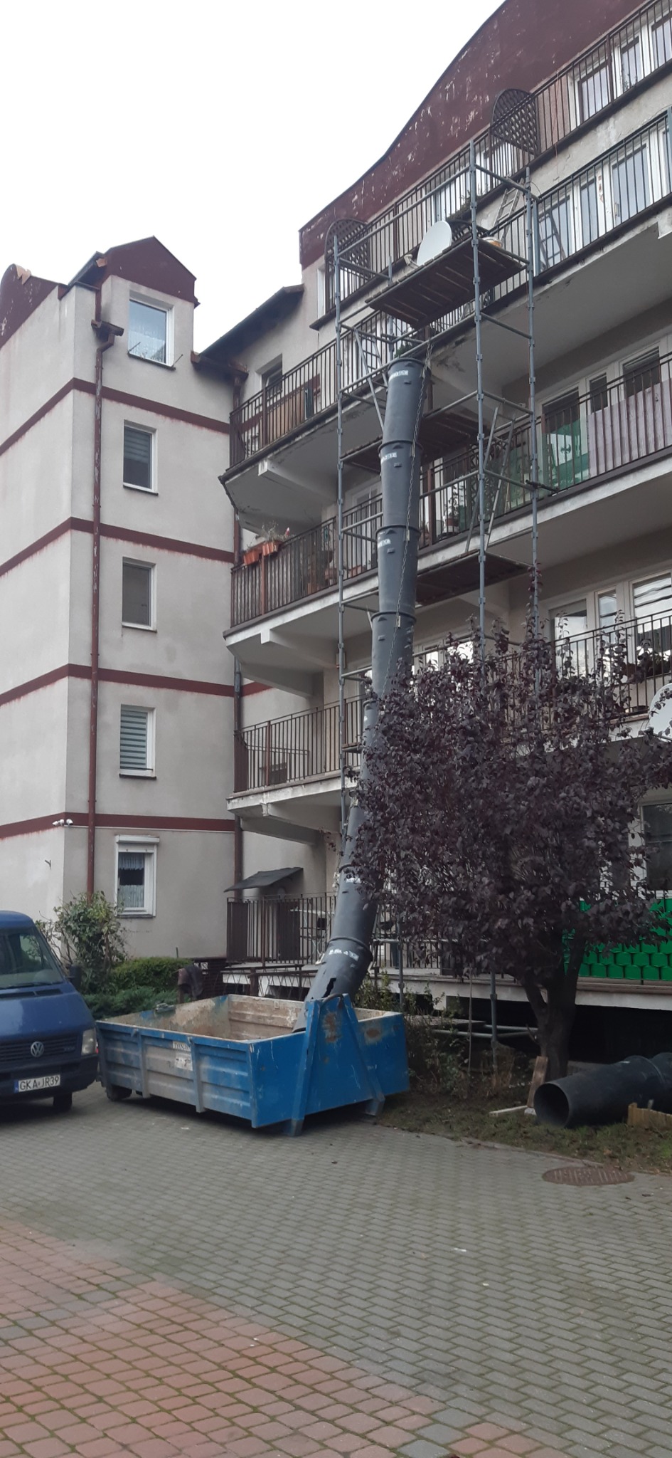 Rynna zsypowa zamontowana na rusztowaniu przy budynku wielorodzinnym, kontener na gruz na dole, widoczny samochód dostawczy.