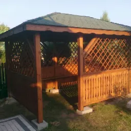 Drewniana altana ogrodowa z zielonym dachem, zdobiona kratownicą, osadzona na betonowych podstawach w otoczeniu zieleni. Widok z zewnątrz.