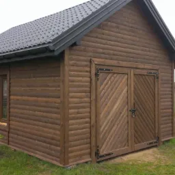 Drewniany domek narzędziowy z bali w Zbuczynie, z dwuskrzydłowymi drzwiami i oknem, na tle zielonego trawnika i ogrodzenia. Ciemna blacha na dachu.