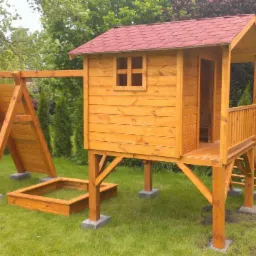 Drewniany plac zabaw z domkiem na palach, huśtawką i piaskownicą w ogrodzie. Czerwony dach domku, naturalne drewno. Zestaw na trawniku, drzewa w tle.