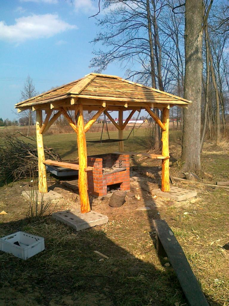 Drewniana altana z ceglanym grillem w ogrodzie. Konstrukcja z bali, dach kryty drewnem, tło stanowią drzewa i łąka.