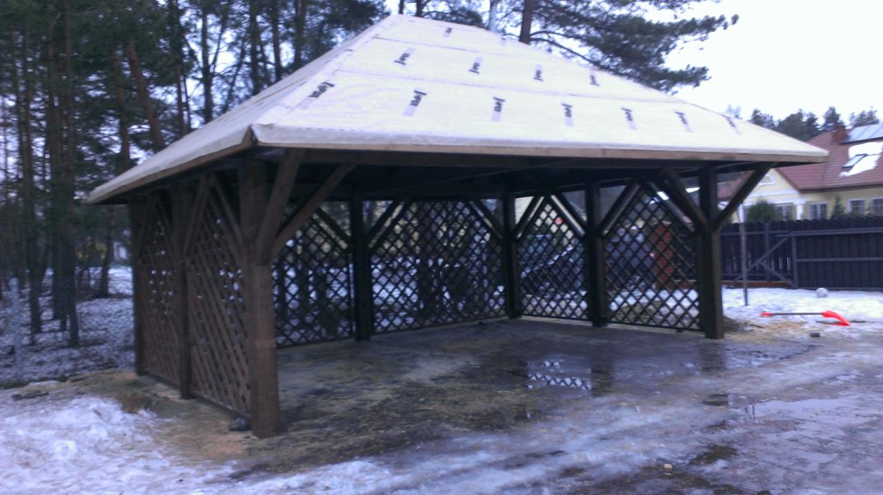 Drewniana altana z kratownicą, przykryta białym materiałem, otoczona lasem i śniegiem, z widocznym fragmentem domu w tle.
