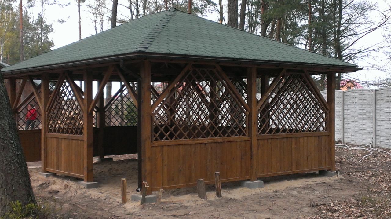 Drewniana altana ogrodowa z zielonym dachem, częściowo osłonięta kratownicą, na tle drzew i betonowego ogrodzenia. Widoczny fragment pnia drzewa po lewej stronie.