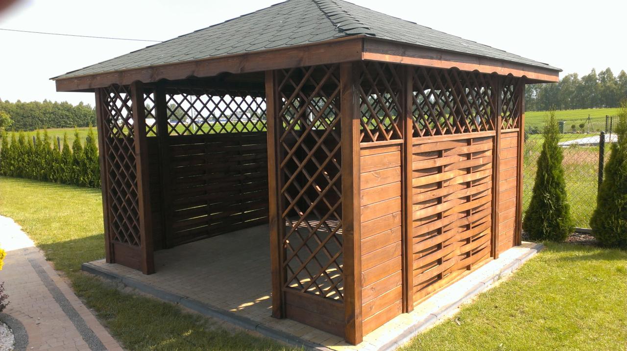 Drewniana altana ogrodowa z ażurowymi ścianami i zielonym dachem, na tle trawnika i żywopłotu. Widoczne detale konstrukcji i wykończenia.