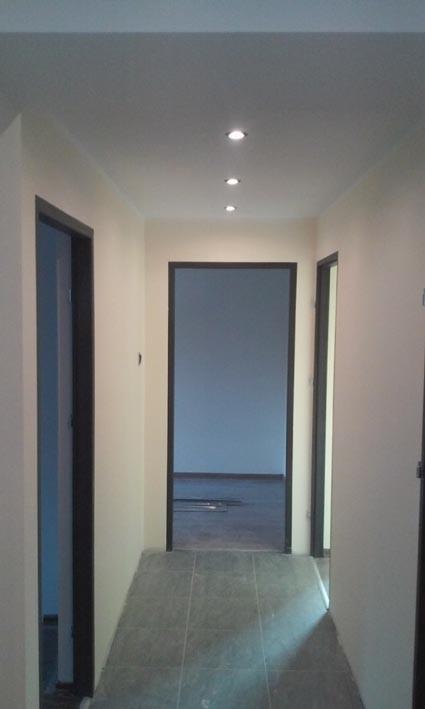 Korytarz po remoncie z szarymi płytkami podłogowymi i trzema parami drzwi z ciemnymi ościeżnicami. Sufit z oświetleniem punktowym wzdłuż korytarza.