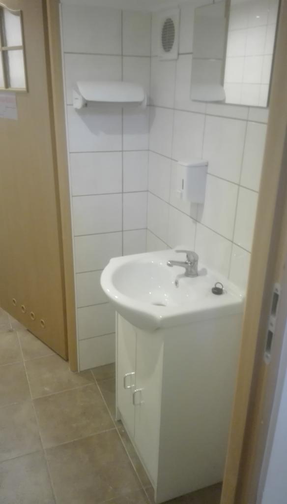 Biała umywalka z szafką w łazience wyłożonej białymi płytkami. Widoczny dozownik mydła, lustro i wentylator. Ujęcie z poziomu podłogi.