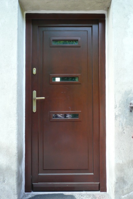 Ciemnobrązowe drzwi zewnętrzne z trzema prostokątnymi przeszkleniami, zamontowane w jasnym, otynkowanym murze. Złota klamka po lewej stronie.