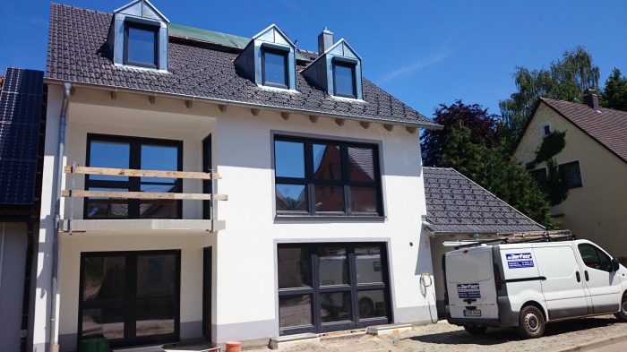 Dwupiętrowy dom z ciemnymi oknami i panelami słonecznymi. Na balkonie widoczne drewniane barierki. Biała elewacja, szary dach i firmowy van na podjeździe.