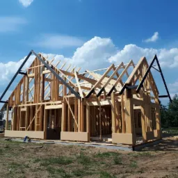 Konstrukcja szkieletowa domu jednorodzinnego w trakcie budowy, widoczne drewniane elementy i czarne metalowe wsporniki dachu, z boku zaparkowany biały bus z logotypem firmy.