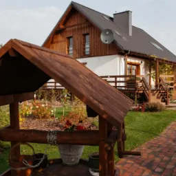 Drewniana studnia z wiaderkami na łańcuchach na pierwszym planie, w tle dom z drewnianą elewacją i zadaszonym tarasem, ceglana ścieżka i zadbany ogród.