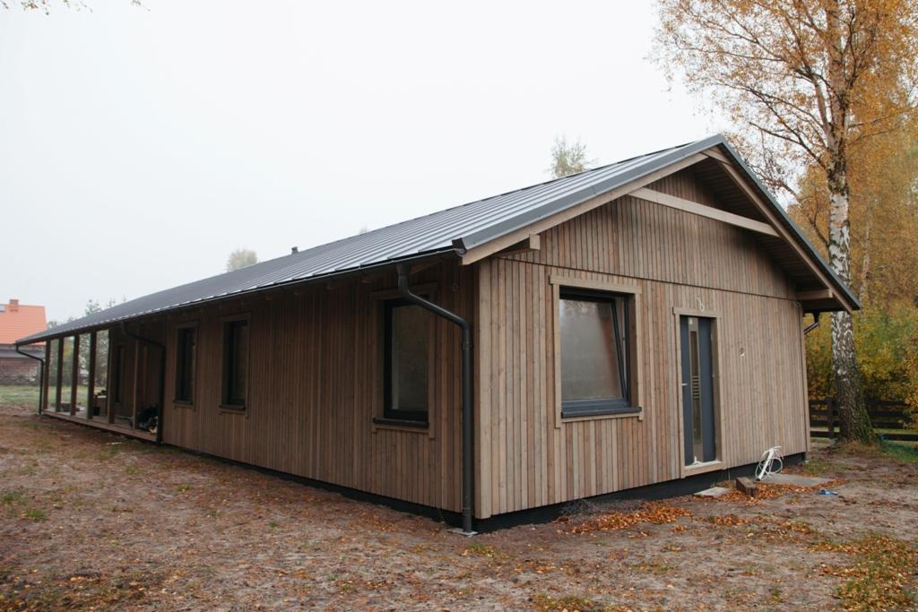 Nowoczesny parterowy dom z jasnego drewna z ciemnym dachem i orynnowaniem, widok z boku na tle jesiennego krajobrazu.