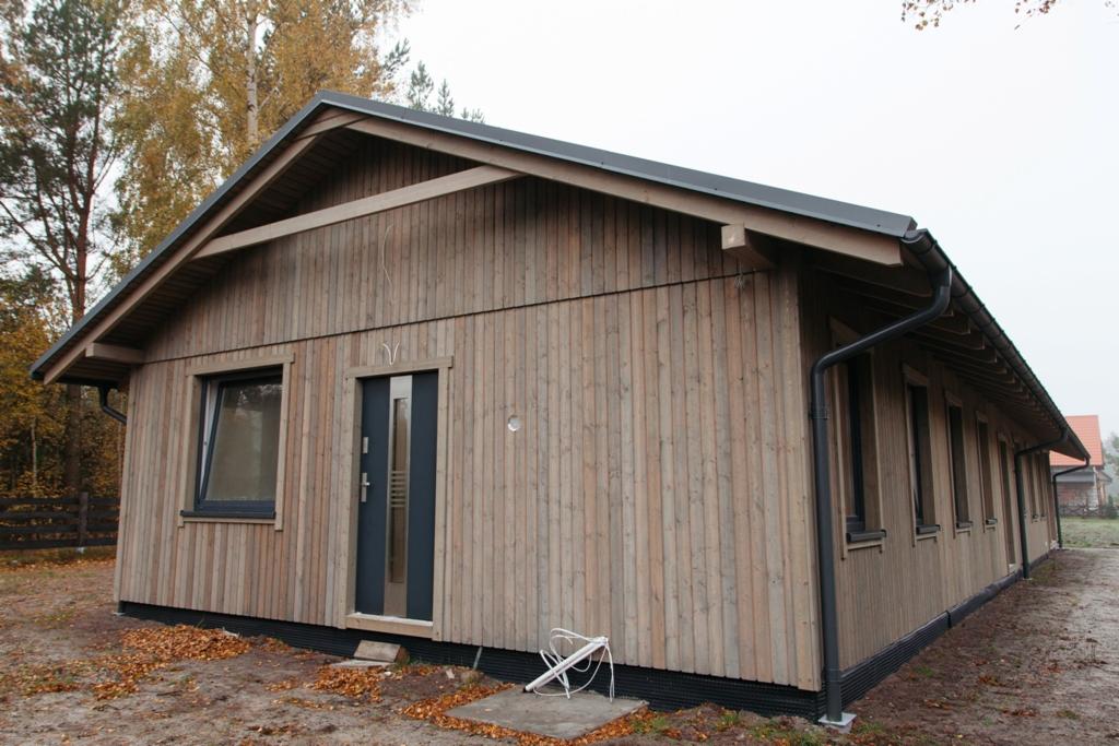 Gotowy budynek z drewnianą elewacją, z widocznymi oknami i drzwiami w ciemnej ramie, z rynnami w kolorze grafitowym, stojący na działce z opadłymi liśćmi, w tle drzewa bez liści.