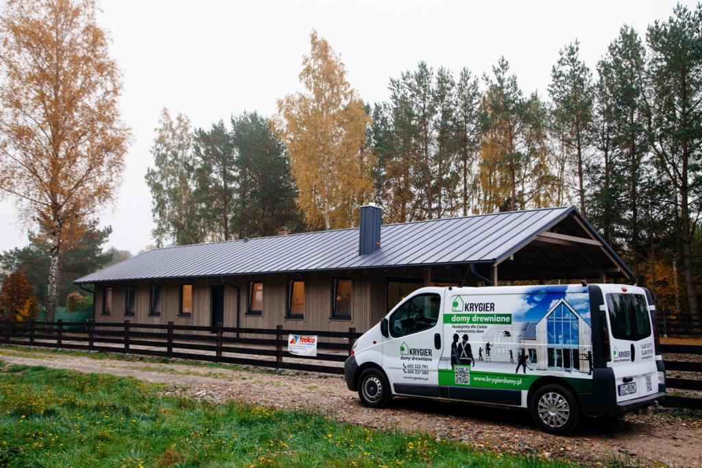 Gotowy dom szkieletowy z szarą blachodachówką, widoczny zza czarnego ogrodzenia i firmowego samochodu dostawczego z reklamą Krygier Domy Drewniane.