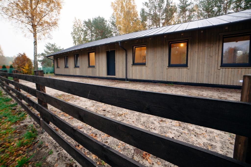 Nowoczesny budynek z jasnego drewna z ciemnymi ramami okiennymi i metalowym dachem, widziany przez czarne, drewniane ogrodzenie w jesiennej scenerii.