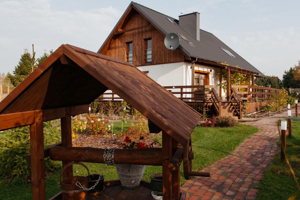 Drewniana studnia z wiaderkami na łańcuchach na pierwszym planie, w tle dom z drewnianą elewacją i zadaszonym tarasem, ceglana ścieżka i zadbany ogród.