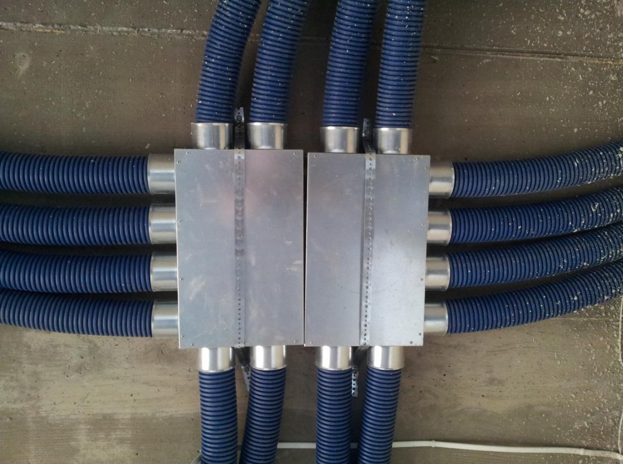 Widok z góry na system rozdzielczy z niebieskimi, karbowanymi przewodami wentylacyjnymi podłączonymi do dwóch metalowych skrzynek rozdzielczych, zamontowanych na szorstkim, betonowym suficie.