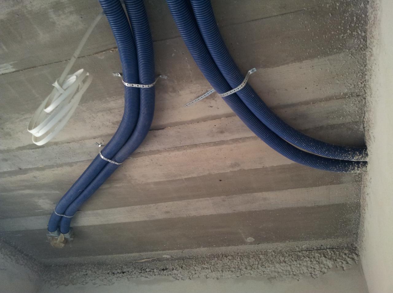 Niebieskie, karbowane rury wentylacyjne przymocowane metalowymi obejmami do surowego betonowego sufitu podczas instalacji.