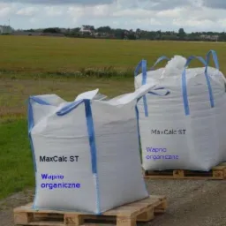 Białe worki typu Big Bag z nawozem wapno organiczne MaxCalc ST na drewnianych paletach, w tle pole i zabudowania.