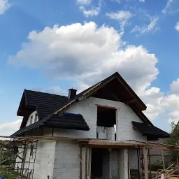 Budowa domu z białych pustaków, widoczny dach z czarnej dachówki i rusztowania. Błękitne niebo z białymi chmurami w tle.