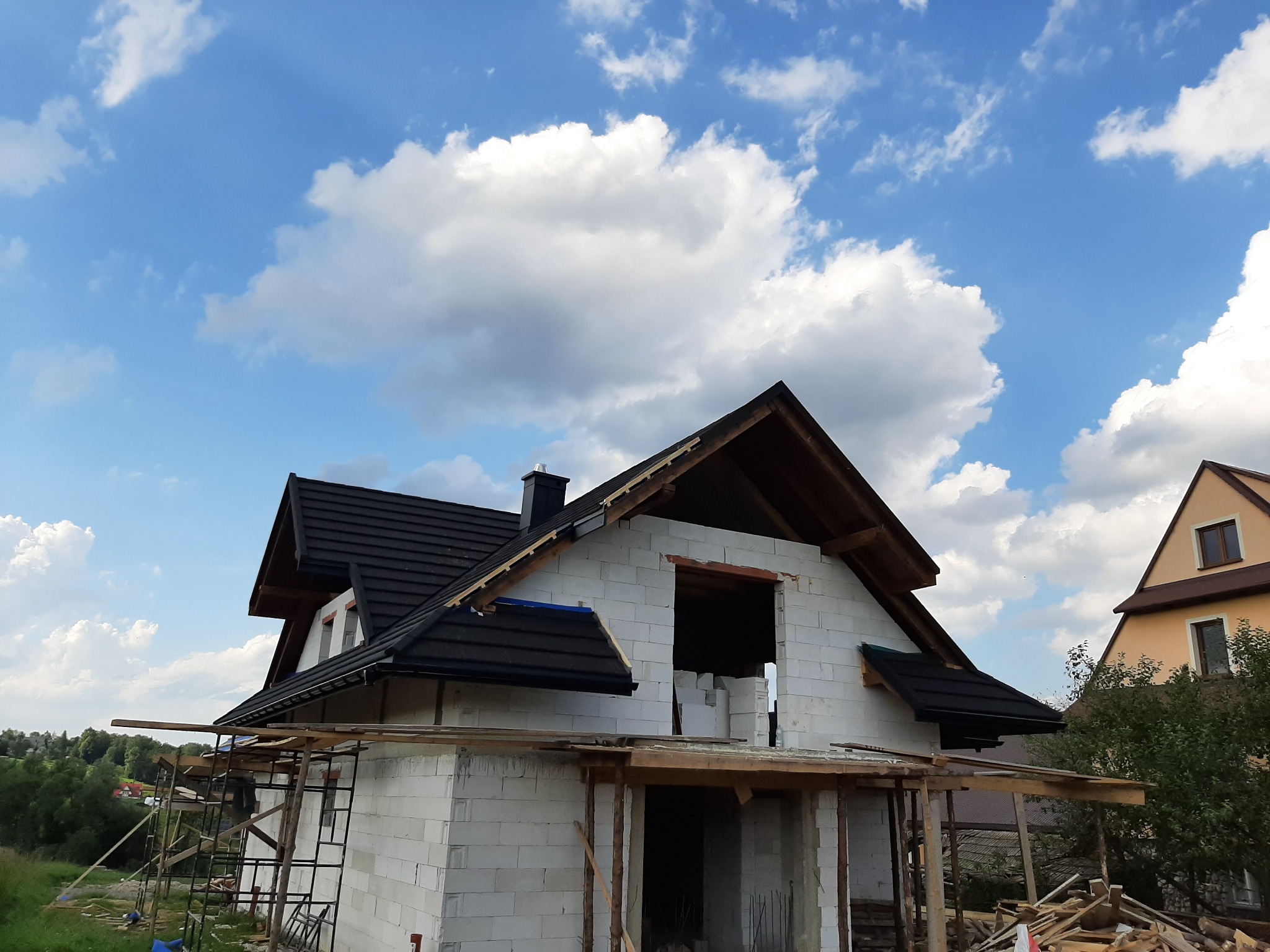 Budowa domu z białych pustaków, widoczny dach z czarnej dachówki i rusztowania. Błękitne niebo z białymi chmurami w tle.
