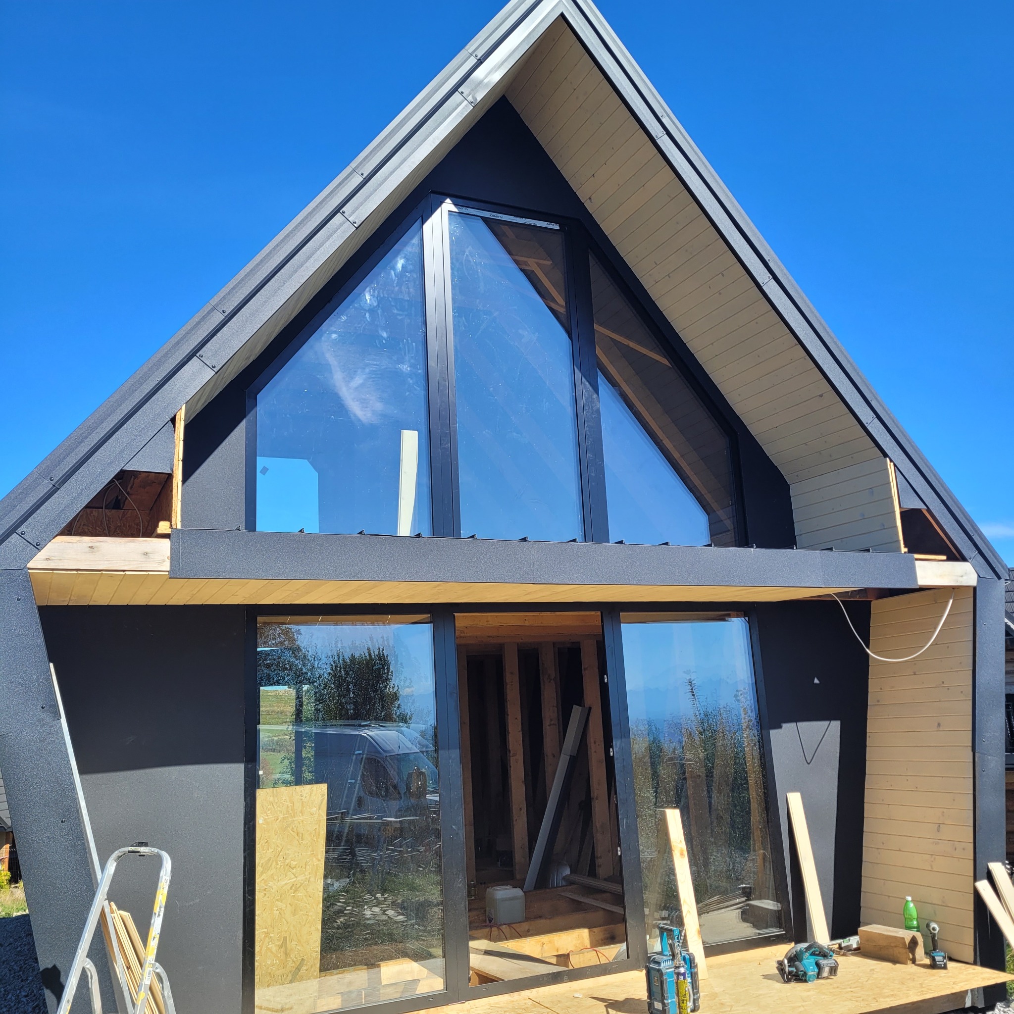 Nowoczesny dom w trakcie budowy z dużym trójkątnym oknem i czarną elewacją. Widoczne narzędzia i materiały budowlane. Jasne, słoneczne niebo w tle.