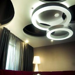 PUH Konkurent Bartłomiej Pawelec - Nowoczesne oświetlenie sufitowe z okrągłymi lampami LED w salonie. Ciemne, dekoracyjne wnęki i szare zasłony w tle. Stylowy wystrój wnętrza.