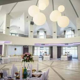 PUH Konkurent Bartłomiej Pawelec - Elegancka sala bankietowa z okrągłym parkietem, stołami z białymi obrusami i kwiatowymi dekoracjami. Białe, kuliste lampy wiszące pod sufitem.