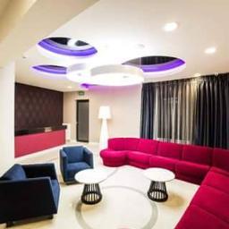 PUH Konkurent Bartłomiej Pawelec - Nowoczesne wnętrze holu z okrągłym sufitem podwieszanym z oświetleniem LED, obszerną sofą w odcieniach różu i ciemnymi zasłonami w tle.