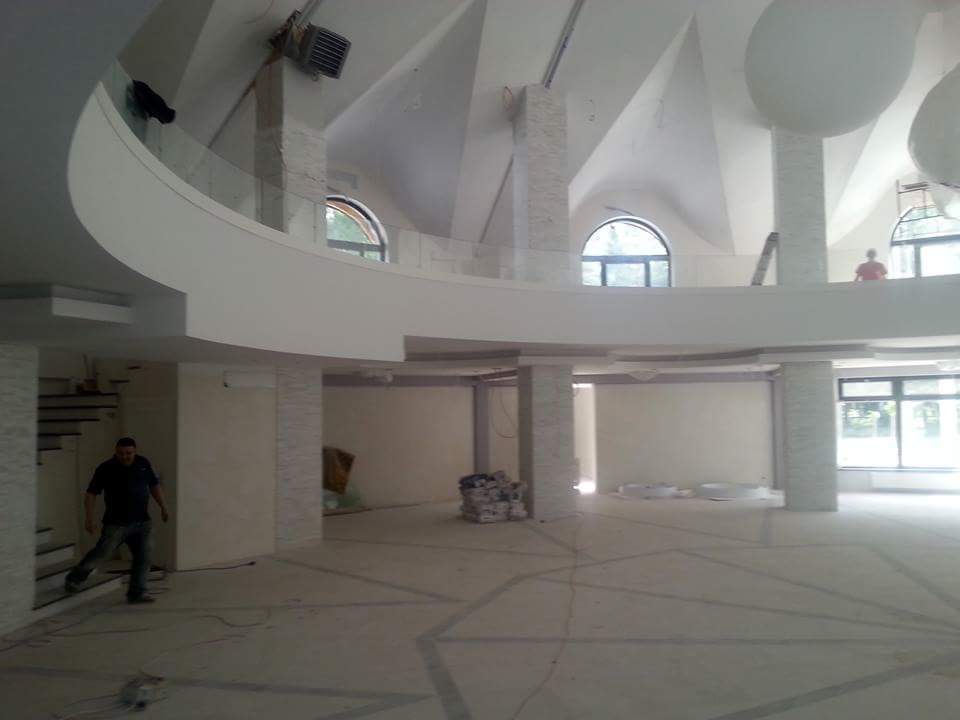 Wnętrze w trakcie remontu: surowe ściany, kolumny z dekoracyjną fakturą, okna łukowe i widoczny zarys wzoru na posadzce. Osoba w tle i na pierwszym planie.