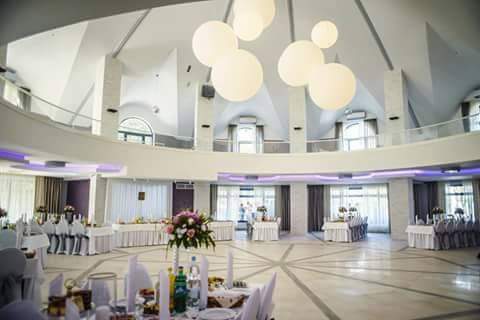 Elegancka sala bankietowa z okrągłym parkietem, stołami z białymi obrusami i kwiatowymi dekoracjami. Białe, kuliste lampy wiszące pod sufitem.