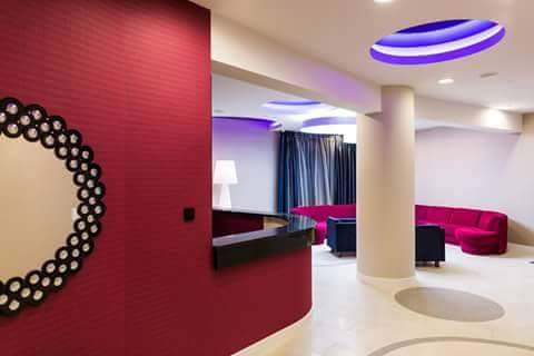 Nowoczesne wnętrze z czerwonymi ścianami, dekoracyjnym lustrem i elementami oświetlenia sufitowego w kolorze fioletowym. Widoczna recepcja i strefa wypoczynkowa.