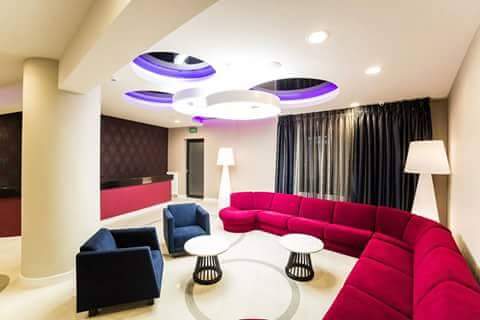 Nowoczesne wnętrze holu z okrągłym sufitem podwieszanym z oświetleniem LED, obszerną sofą w odcieniach różu i ciemnymi zasłonami w tle.