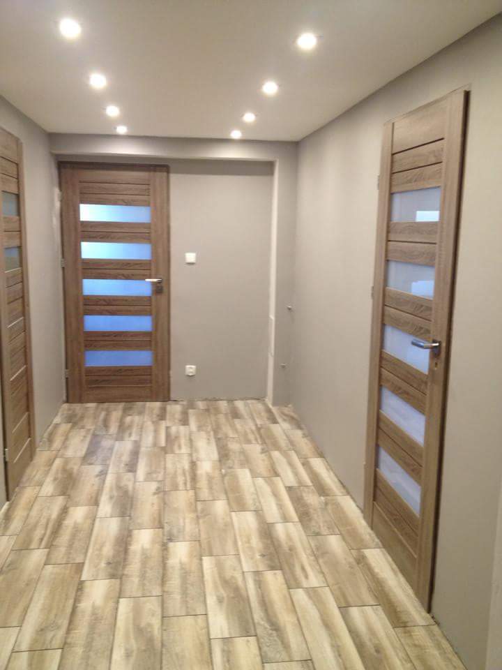 Korytarz z trzema drewnianymi drzwiami z przeszkleniami, oświetlony punktowo. Podłoga wyłożona płytkami imitującymi drewno. Ściany w odcieniach szarości.