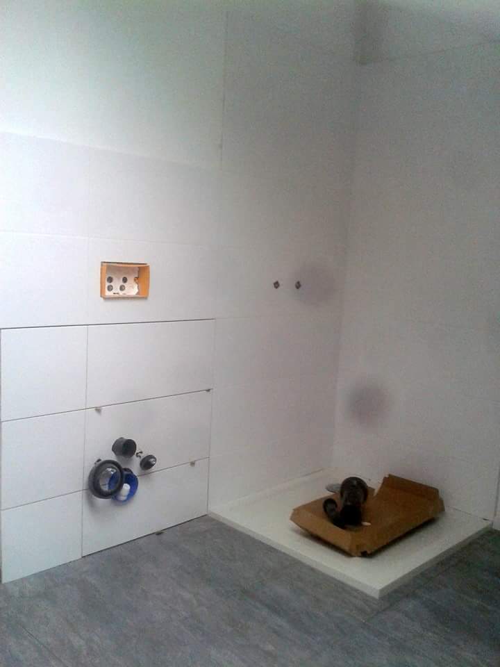 Remont łazienki: białe płytki ścienne, szare podłogowe, zamontowane rury odpływowe pod umywalkę i przygotowany brodzik z odpływem. Otwór na gniazdko elektryczne.