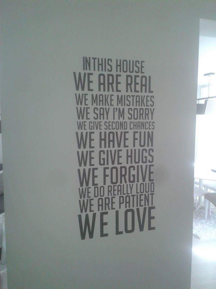 Ściana z dekoracyjnym napisem w szarym kolorze: 'In this house we are real...we love'. W tle widoczne jasne wnętrze z białymi meblami i oświetleniem.
