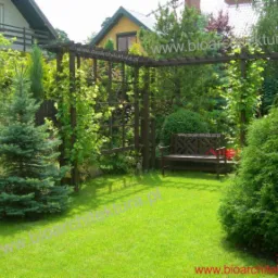 Zadbany ogród z zielonym trawnikiem, pergola z pnączami, ozdobne krzewy i drzewka iglaste, drewniana ławka w tle.