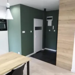 Wnętrze po remoncie: ciemnozielona ściana, jasna podłoga, drewniana okładzina ściany i czarna podłoga w przedpokoju.