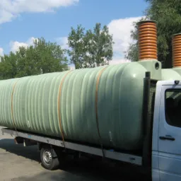 Zielony zbiornik z dwoma kominami wentylacyjnymi, zabezpieczony pasami, przewożony na platformie samochodu dostawczego w słoneczny dzień.