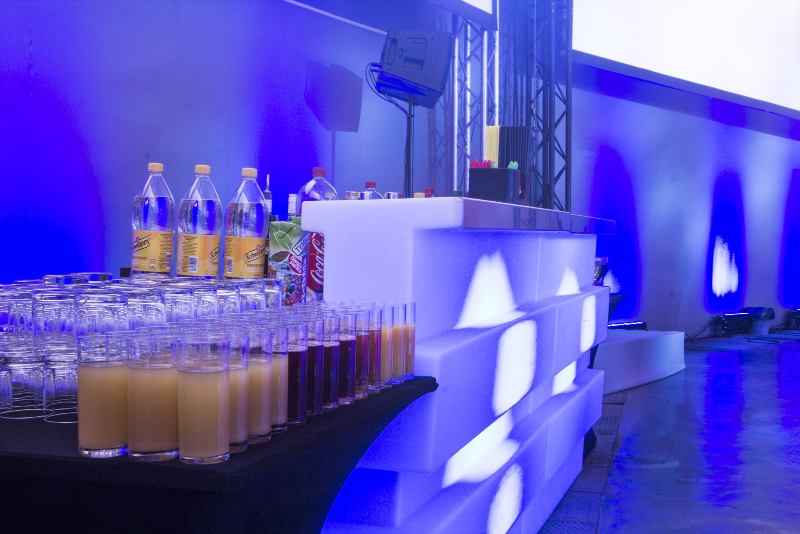 Podświetlony bar z napojami i szklankami, przygotowany na imprezę. W tle widoczne oświetlenie sceniczne i konstrukcja kratownicowa. Dominują odcienie błękitu.