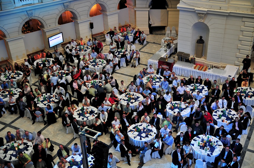 Widok z góry na salę bankietową z okrągłymi stołami, gośćmi w formalnych strojach i bufetem. Uwagę zwraca klasyczna architektura wnętrza.