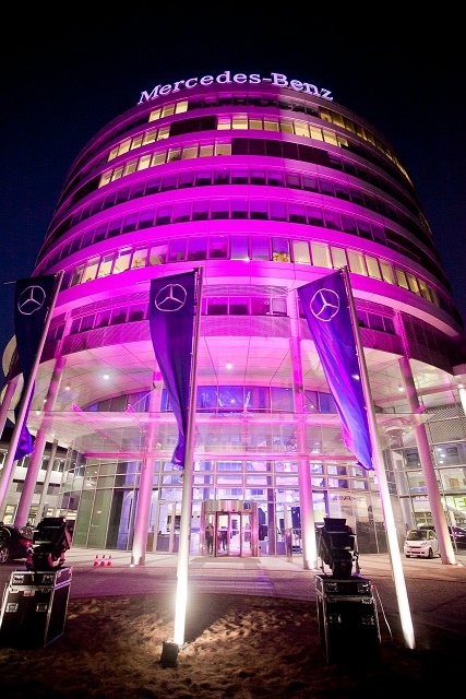Okrągły budynek Mercedes-Benz, nocą podświetlony na różowo, z trzema flagami z logo marki przed wejściem. Widoczne nagłośnienie i oświetlenie eventowe.