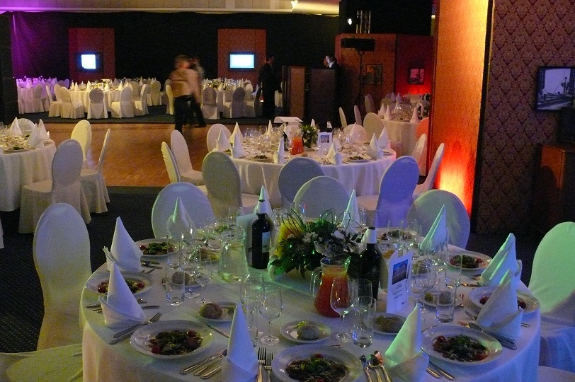 Uroczysta sala bankietowa z okrągłymi stołami, nakryta białą zastawą i dekoracjami kwiatowymi, przygotowana na przyjęcie. W tle widoczni goście.