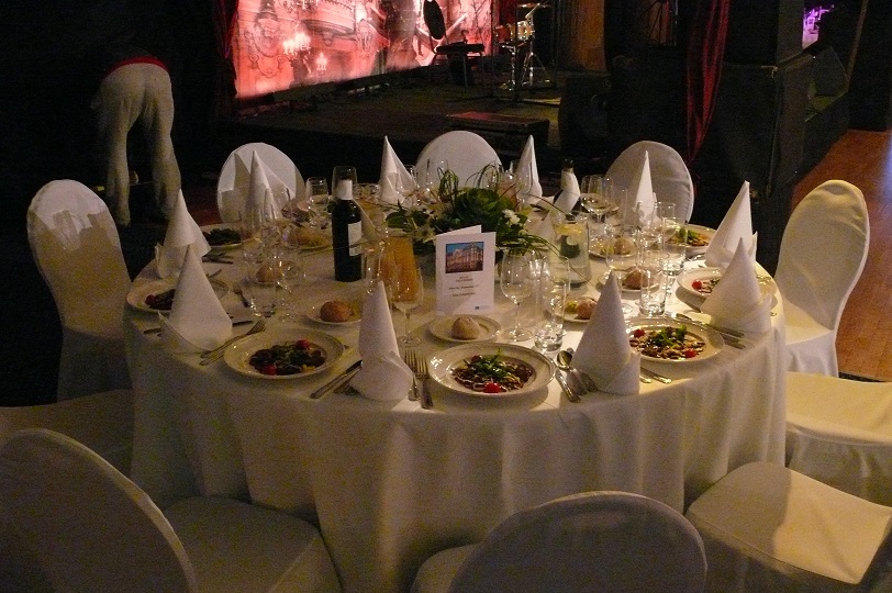Uroczysty stół z białym obrusem, nakryty dla gości, z dekoracjami kwiatowymi i zastawą, w tle scena z nagłośnieniem, oświetleniem i osobą w kostiumie.