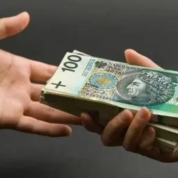 Dwie dłonie w garniturach wymieniają się plikiem banknotów 100 zł na ciemnym tle, sugerując transakcję finansową lub pożyczkę.