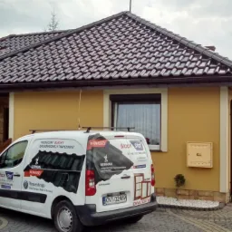 Dom z brązową dachówką modułową, widoczny z podjazdu. Na pierwszym planie firmowy bus z reklamą producenta blach dachowych i okien. Część dachu zakryta folią.