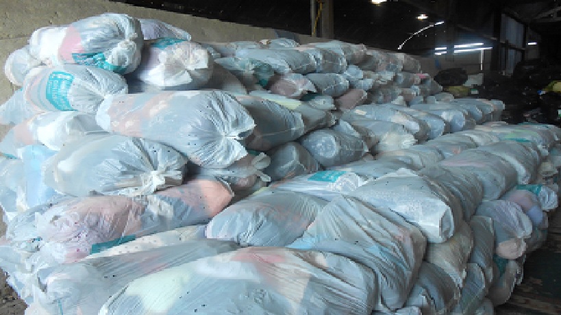 Stos worków z odzieżą używaną, zapakowanych w przezroczyste plastikowe torby, ułożonych warstwami w magazynie lub hurtowni. Widoczne zarysy ubrań wewnątrz.