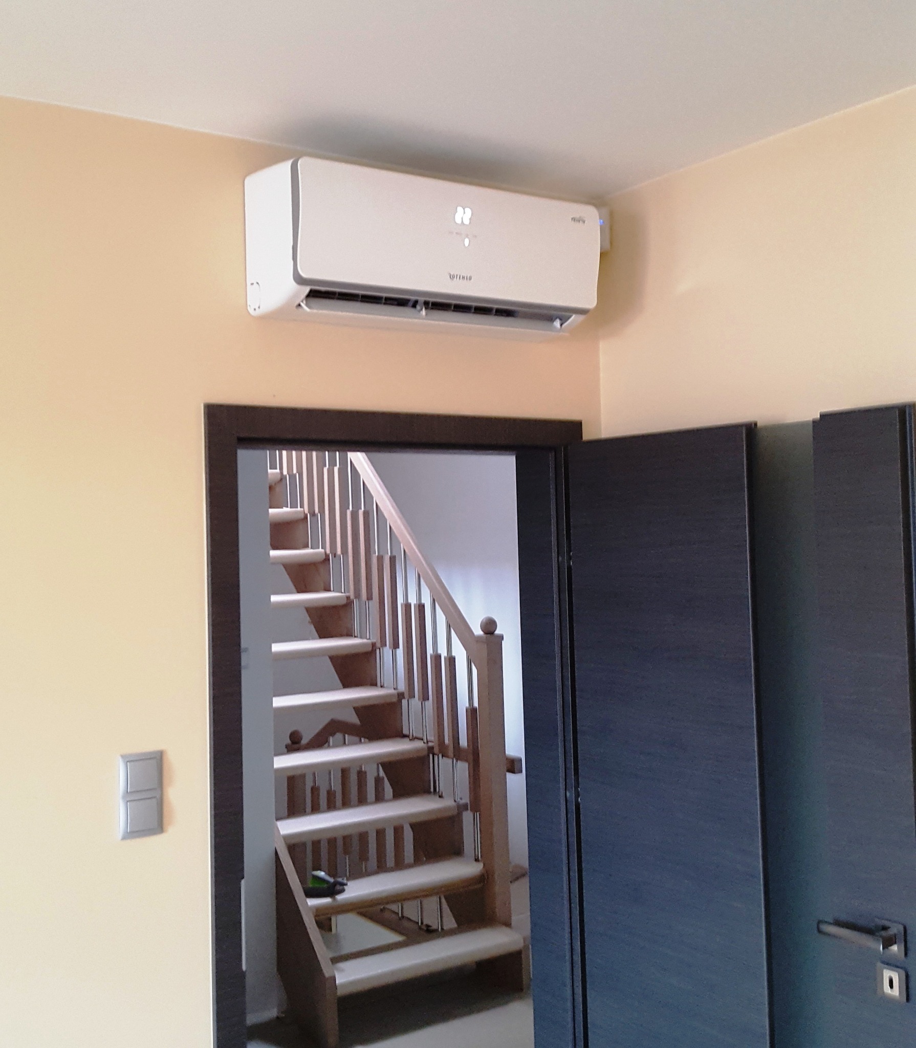 Biała jednostka klimatyzacji zamontowana na ścianie nad drzwiami, widoczna drewniana klatka schodowa w tle.