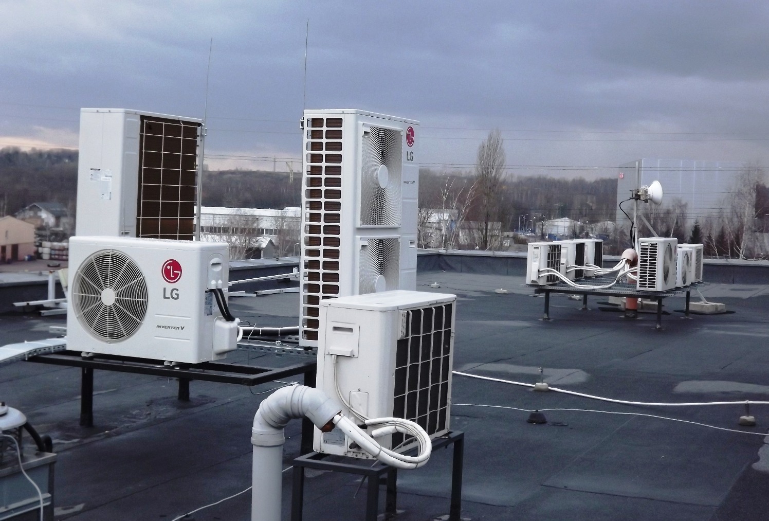 Instalacja kilku zewnętrznych jednostek klimatyzacyjnych LG na płaskim dachu budynku komercyjnego, widoczne przewody i rury odprowadzające skropliny, tło stanowią budynki przemysłowe i drzewa...