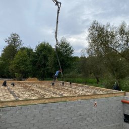 Zielpol 2.0 Dariusz Zieliński - Wylewka fundamentu domu z betonomieszarki. Widoczne zbrojenie, szalunek i ściany fundamentowe z bloczków. W tle drzewa i pochmurne niebo.